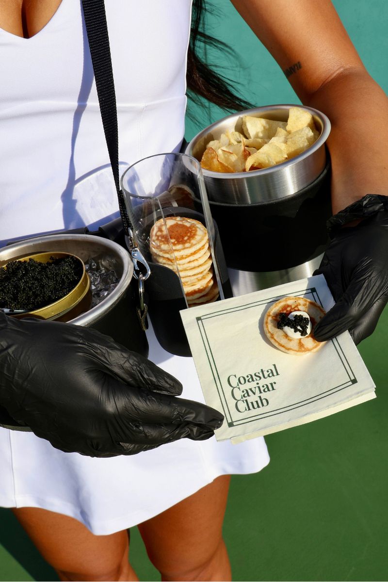 Close-up of the caviar serving belt: a tin of caviar on ice, a stack of blini, kettle chips and a blini topped with caviar on a Coastal Caviar Club napkin