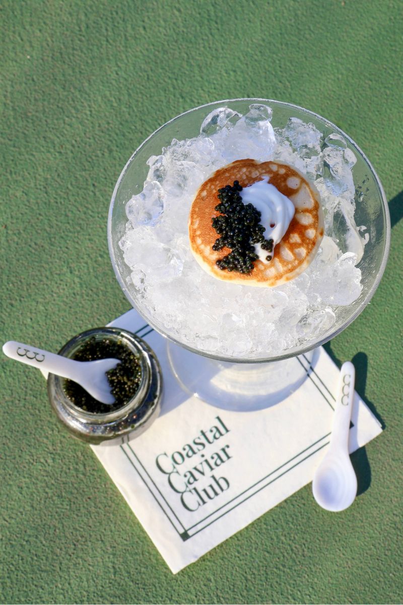 A blini topped with crème fraîche and caviar on crushed ice in a glass coupe, beside a jar of caviar, two club spoons and a Coastal Caviar Club napkin