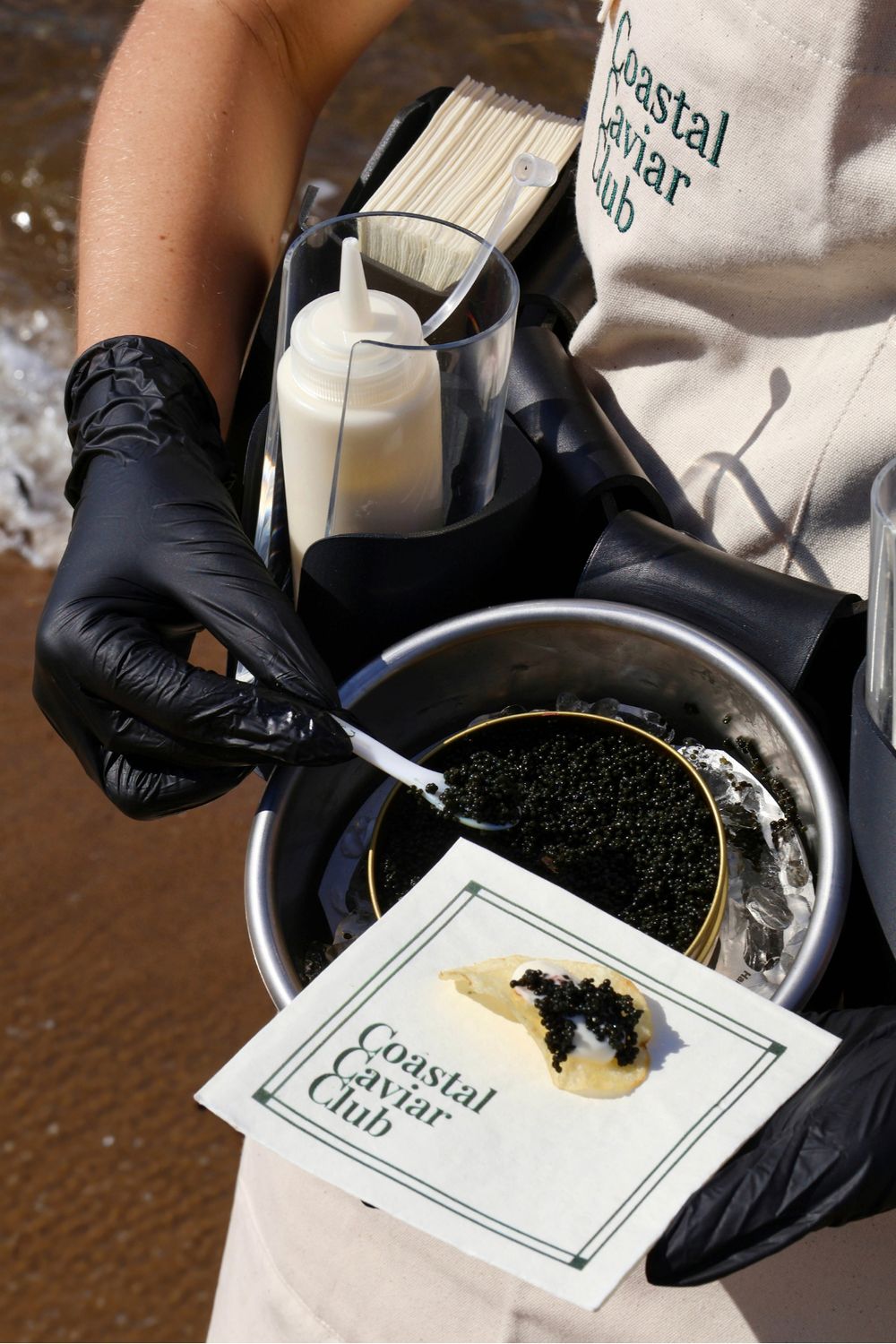 A Caviar Host at the water's edge spooning caviar from a tin on ice, with a blini-style chip of caviar and crème fraîche on a Coastal Caviar Club napkin